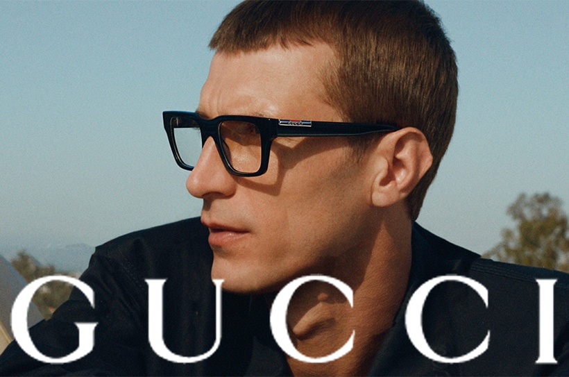 Zijaanzicht van een man met zwarte bril en minimalistische jas, \'GUCCI\' onderaan in beeld