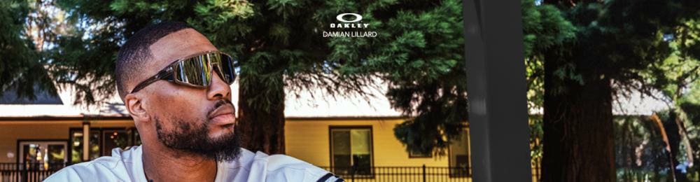 Portret van Damian Lillard, een man met een baard en zonnebril, zittend in de schaduw van bomen, gekleed in een licht sportshirt. Het Oakley-logo is zichtbaar boven hem.