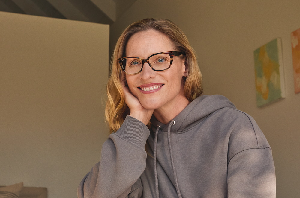 Een glimlachende vrouw met een bril en een grijze hoodie, zittend in een goed verlichte kamer met abstracte schilderijen.