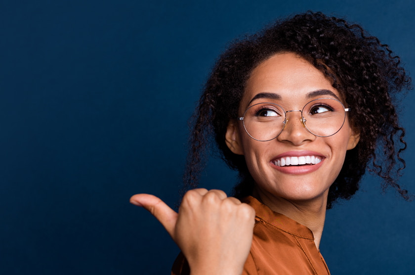 Glimlachende vrouw met een ronde metalen bril, die met haar duim over haar schouder wijst. Ze draagt een koperkleurige blouse en de achtergrond is effen blauw.