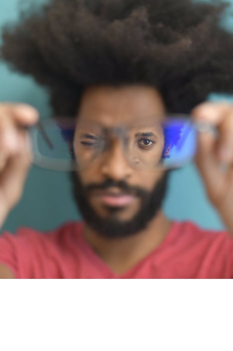 Een man met krullend afrohaar en een baard houdt een bril met blauwlichtfilter voor zich terwijl hij door de glazen kijkt. Hij draagt een rood T-shirt en staat voor een wazige turquoise achtergrond.
